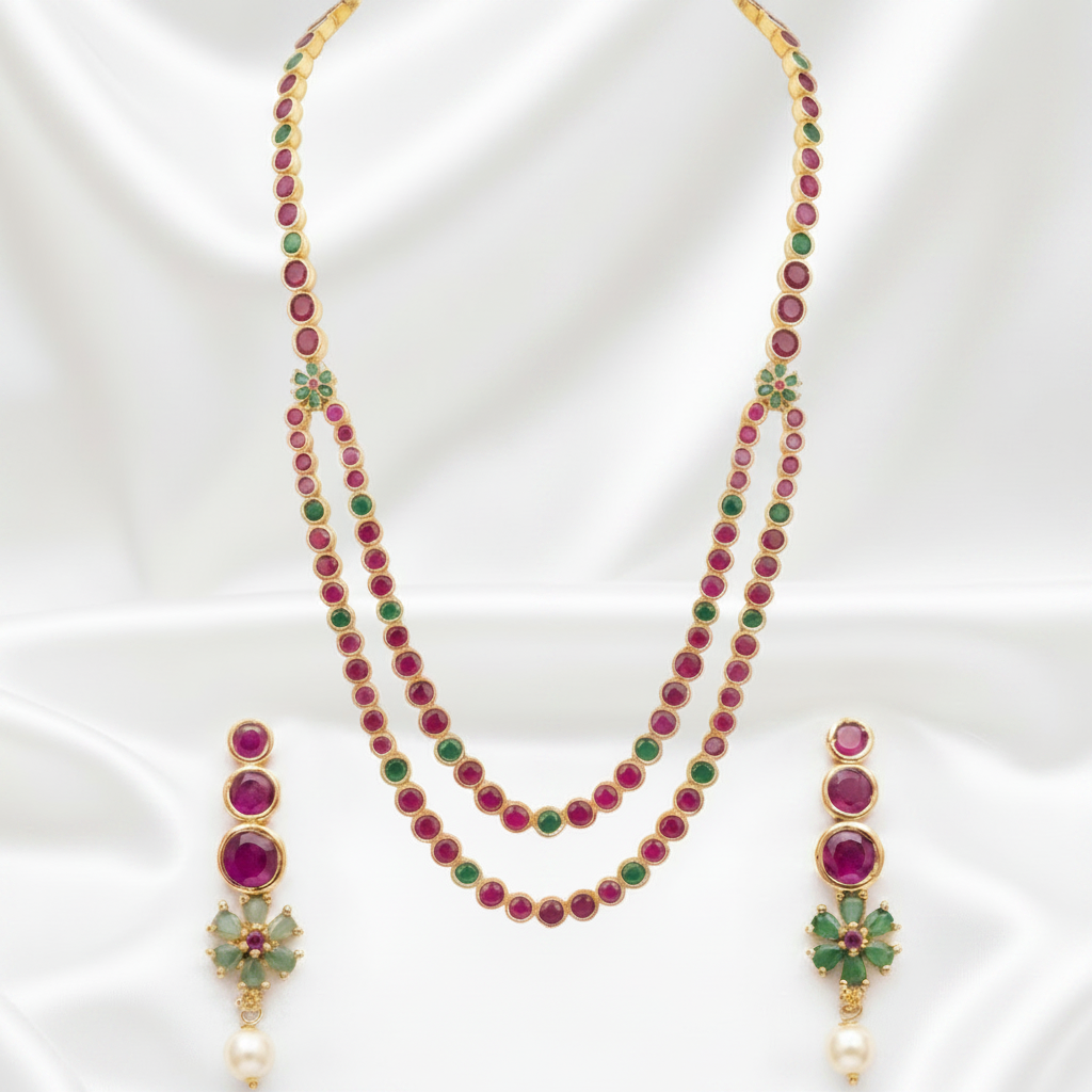 Gold-plated necklace and matching drop earrings set with round magenta-red and green stones, displayed on a white satin background.