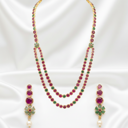 Gold-plated necklace and matching drop earrings set with round magenta-red and green stones, displayed on a white satin background.