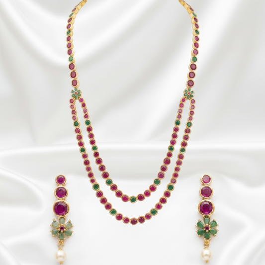 Gold-plated necklace and matching drop earrings set with round magenta-red and green stones, displayed on a white satin background.