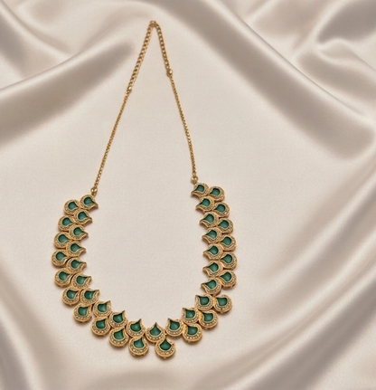 A gold-toned statement necklace featuring a curved, scalloped arrangement of green stone leaf or petal motifs, displayed on draped cream satin fabric.